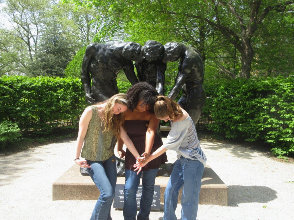 Three students are imitating the statue behind them by leaning over and putting their hands in the middle of them.