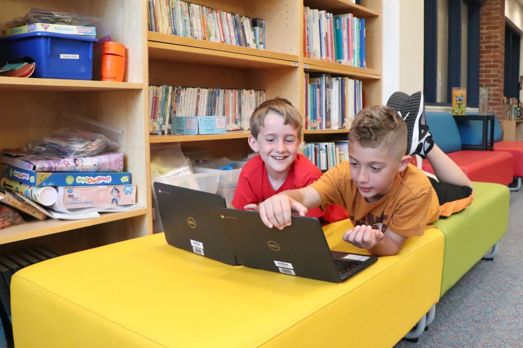 Two students lay on ottomans and look at laptops used for learning in the Scotchtown Avenue School library.