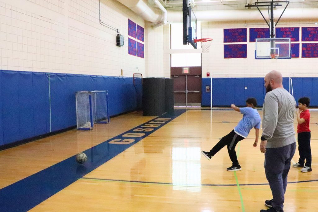 A student kicks a soccer ball at a wall while practicing for an event. There is another student standing in line behind him. Coach Chales Doyle is nearby watching the practice.
