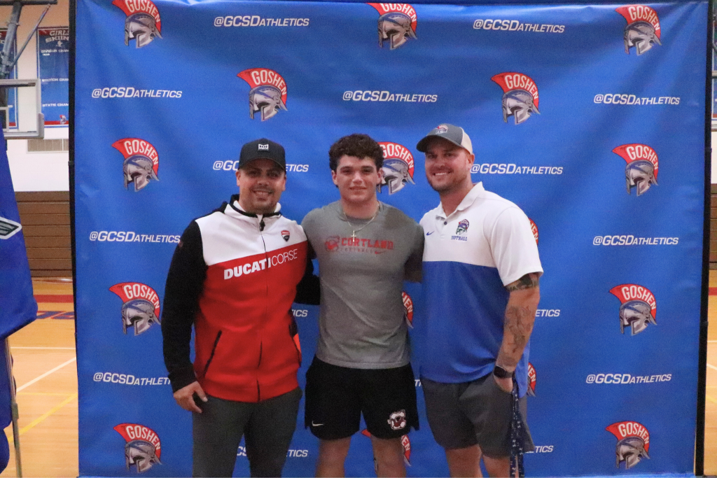 Vincenzo Pupo smiles for a photo in front of the Goshen background. His coaches are on either side of him.
