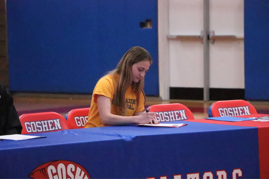 Olivia Grob is signing a letter on a Gladiator table with Goshen chairs next to her. She is wearing a yellow shirt of her college.