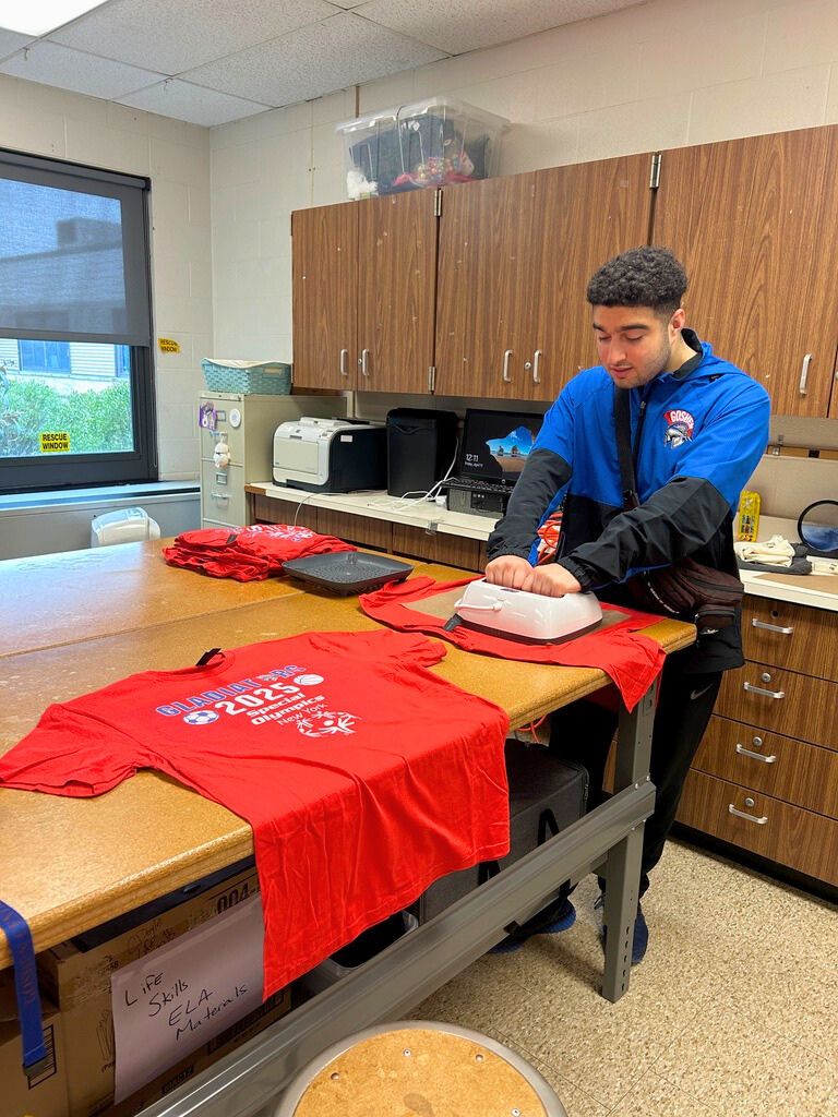 A student is pressing a logo onto a shirt with other shirts on the table next to him. The shirts are red and have the year of the Special Olympics on the table.