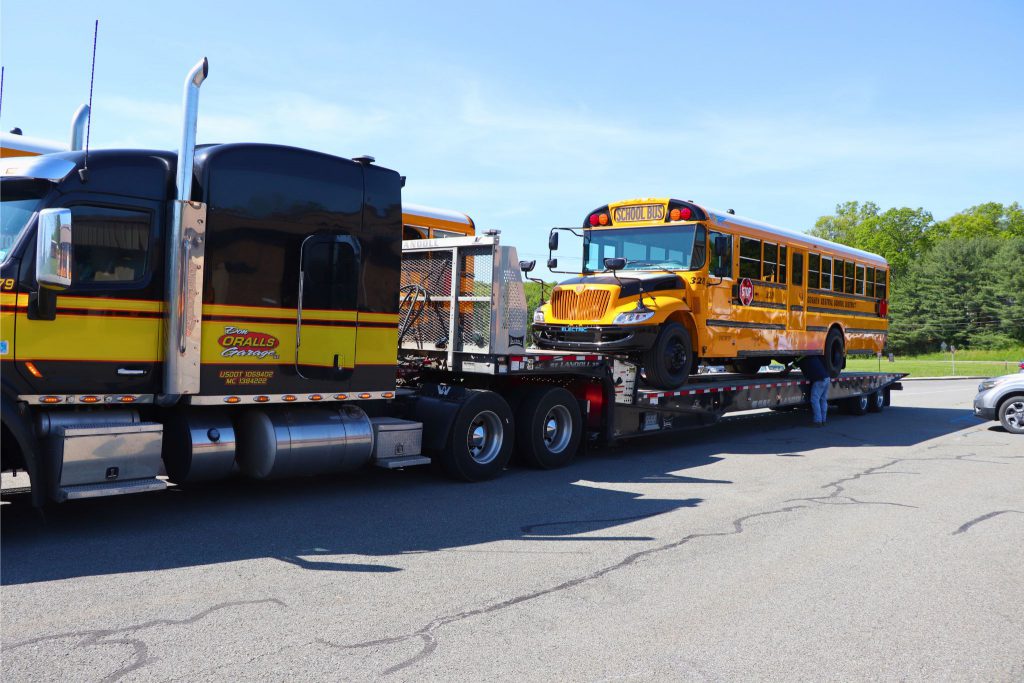 An electric bus is being delivered on a large platform. The bus is new and is shiny in the Goshen transportation parking lot.