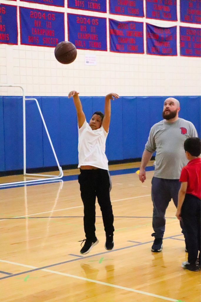 A student shoots a basketball with Coach Charles Doyle nearby watching. There is another student standing on the court as well.