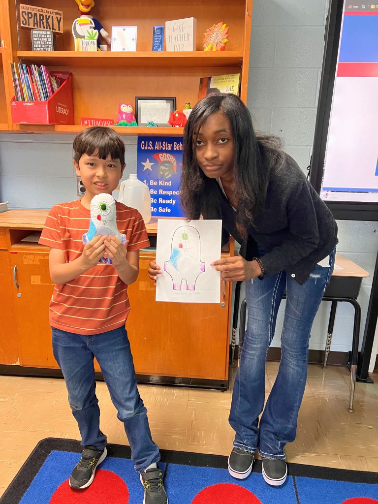 A third grade student stands to the left of a high school student. The young student is holding the sewn monster from the drawing that the older student is holding. The monster is white and has one green eye, with a little red and some blue on it's body as well.