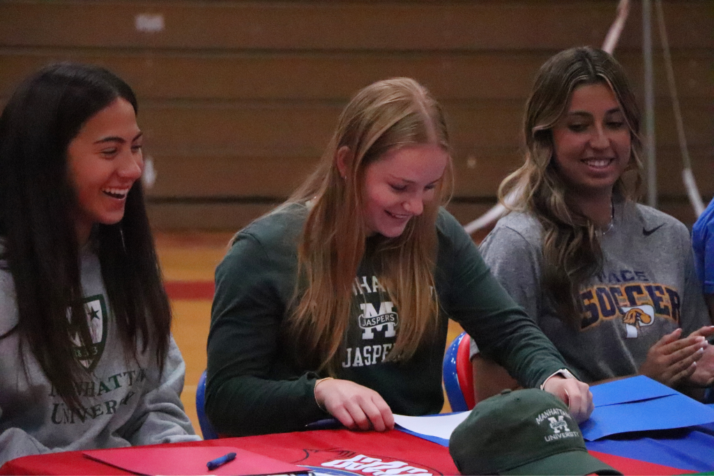 Emma Duffy reaches for her letter to sign on a table with a Goshen logo. Two players are smiling and sitting next to her. There is a Manhattan hat in front of her and she is wearing a Manhattan Jasper shirt.