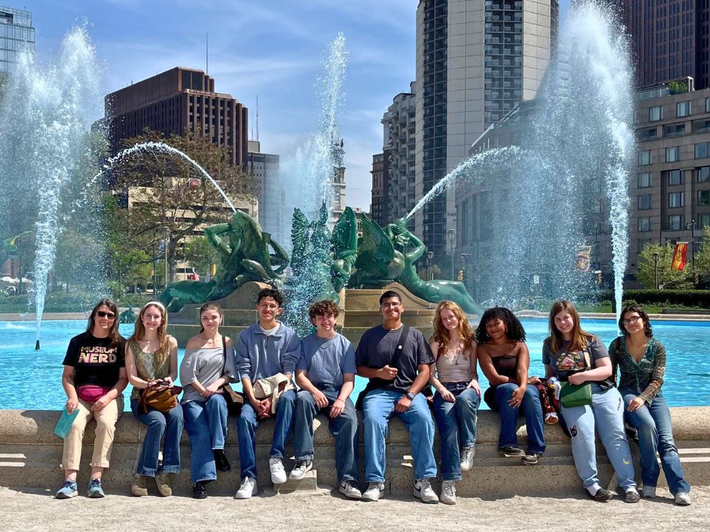 Students are sitting in front of a pool with fountains and taking a group picture.