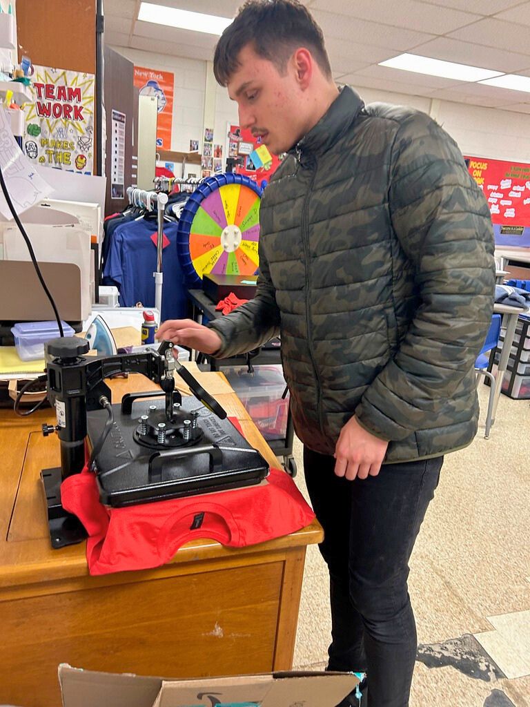 A student is standing next to a machine that is pressing the logo for the Special Olympics onto a red shirt.
