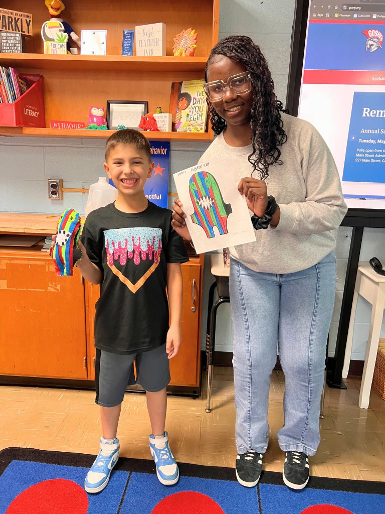 A third grade student stands to the left of a high school student. The young student is holding the sewn monster from the drawing that the older student is holding. The monster is multicolored with green, red and blue and a big mouth near the center of it's body.