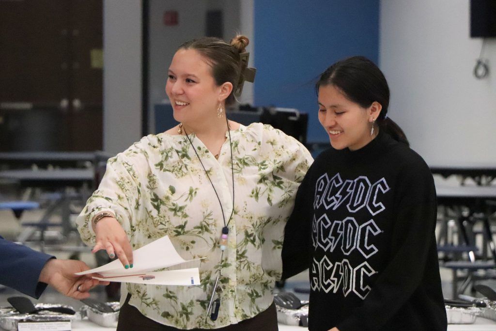 A teacher smiles while reaching out for a paper with a student in the cafeteria.