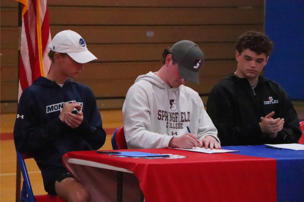 Colin Butryn is signing a paper in a Springfield hat and sweater. There are two athletes next to him clapping as he signs the paper.