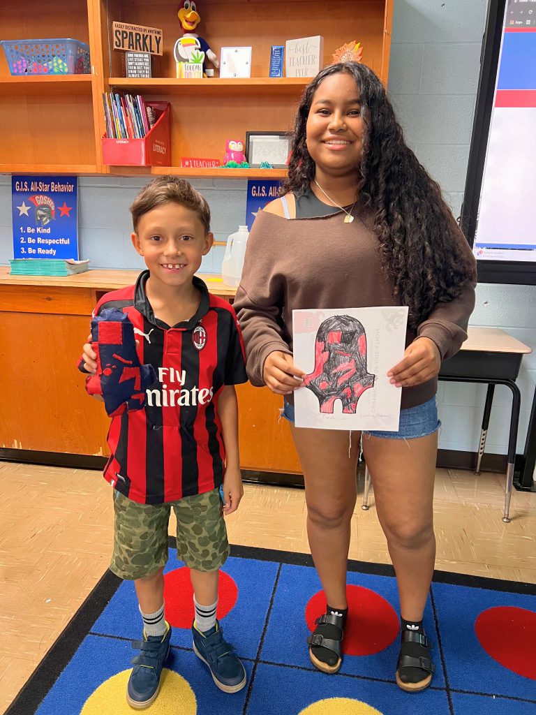 A third grade student stands to the left of a high school student. The young student is holding the sewn monster from the drawing that the older student is holding. The monster is red and dark blue.