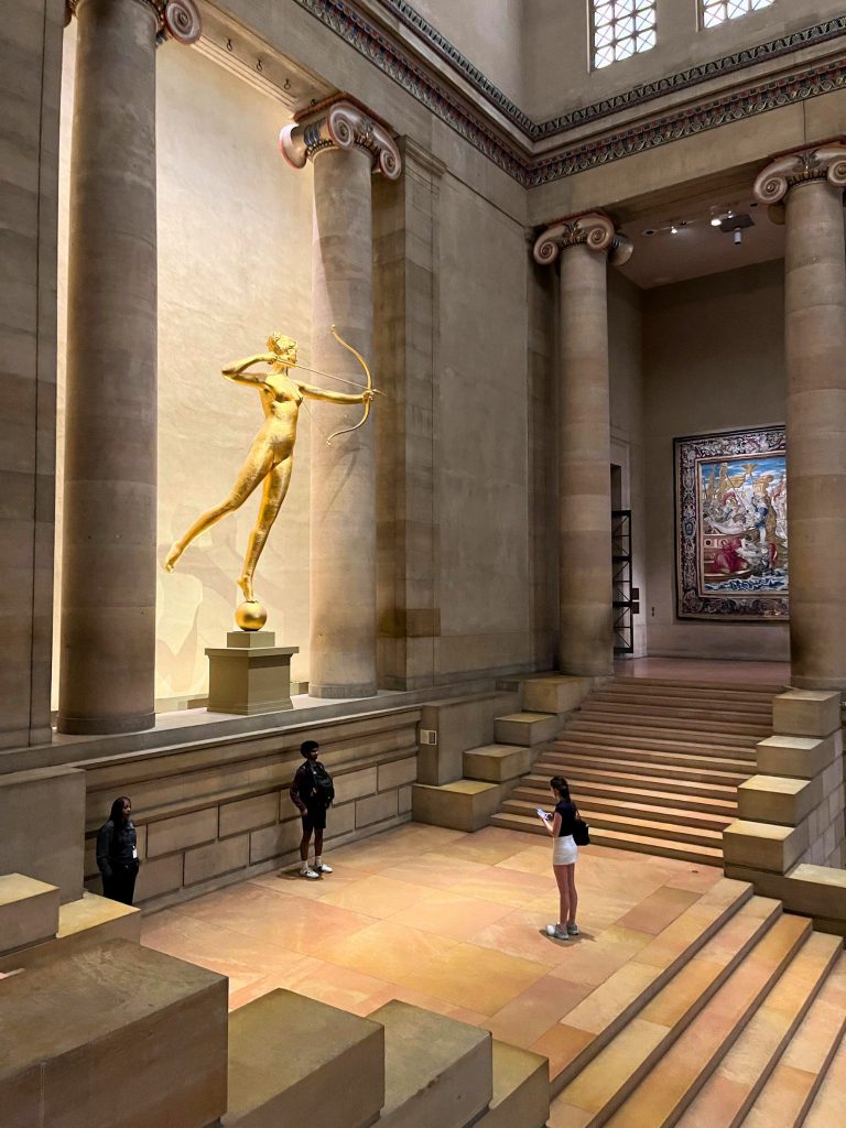 Students pose for a photo under a gold person with a bow and arrow in a large open museum.