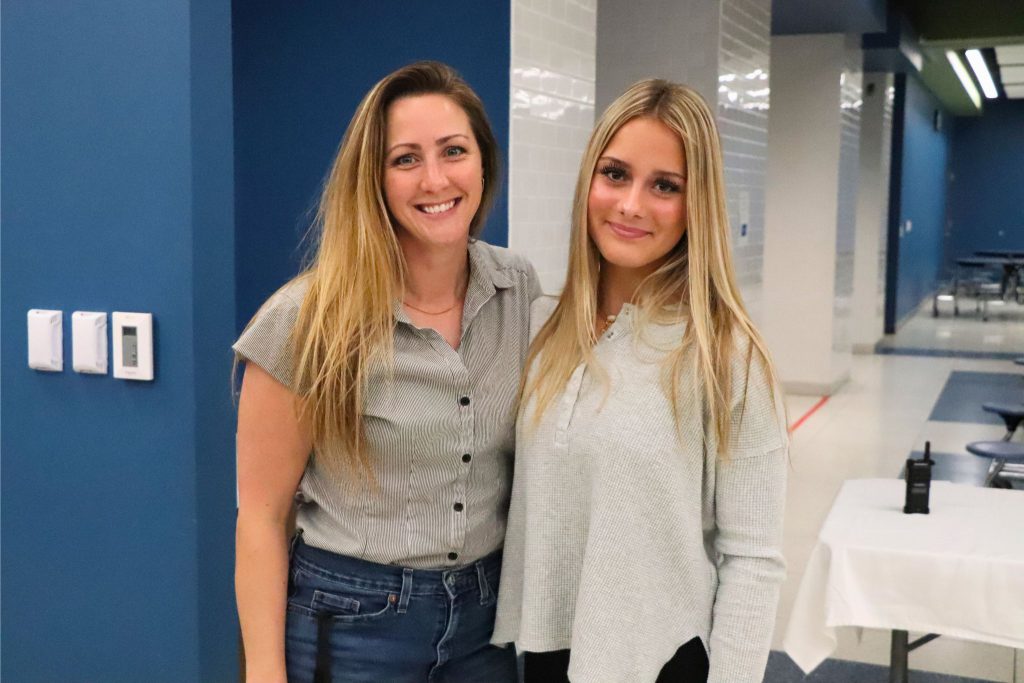 A teacher and a student stand next to each other in the cafeteria. They are both looking at the camera and smiling for a photo.