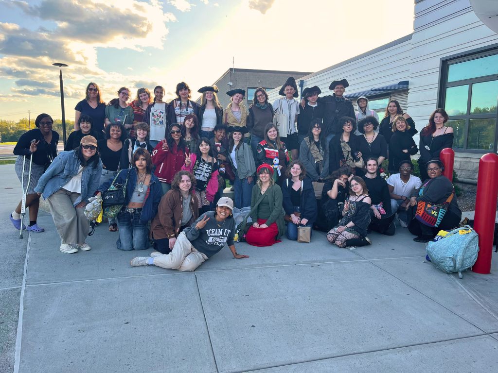 Students take a picture together in front of Goshen High School once they've returned from their trip. They are all wearing clothes from the trip and standing near each other in front of the school entrance.