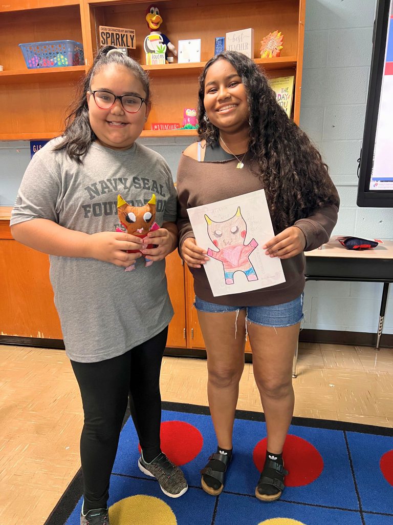 A third grade student stands to the left of a high school student. The young student is holding the sewn monster from the drawing that the older student is holding. The monster is tan, red and blue and has pointy ears off the top of its head.
