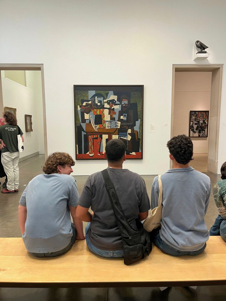 Three students are sitting and looking at a painting on the wall. The photo is taken from behind their heads.