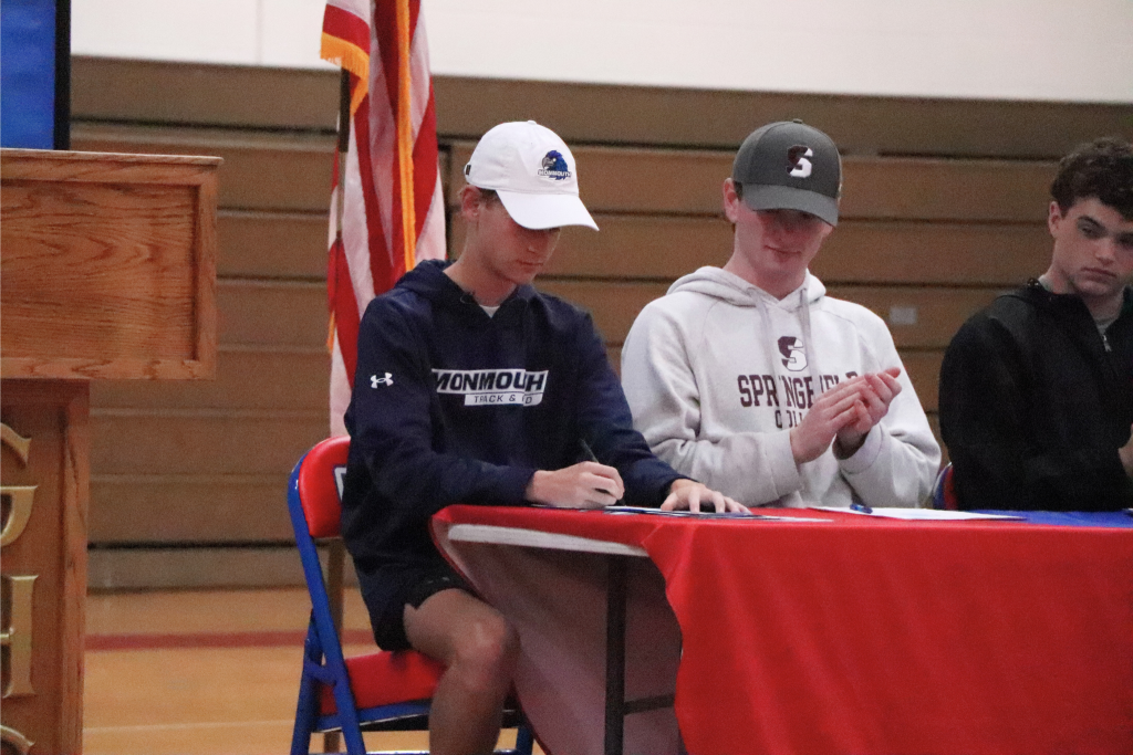 Noah Klugman is wearing a Monmouth sweater and hat and signing a paper with an athlete next to him clapping.
