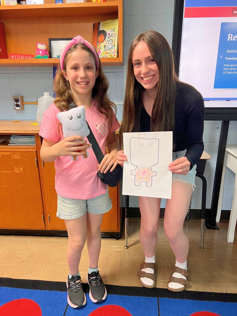 A third grade student stands to the left of a high school student. The young student is holding the sewn monster from the drawing that the older student is holding. The monster has a gray head and big eyes and is smiling.