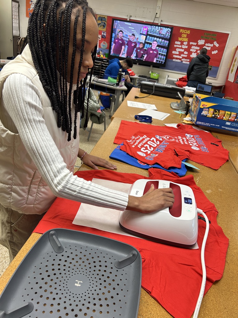 A student is holding a machine over a shirt and is pressing a logo onto a red shirt for the Elementary Special Olympics.