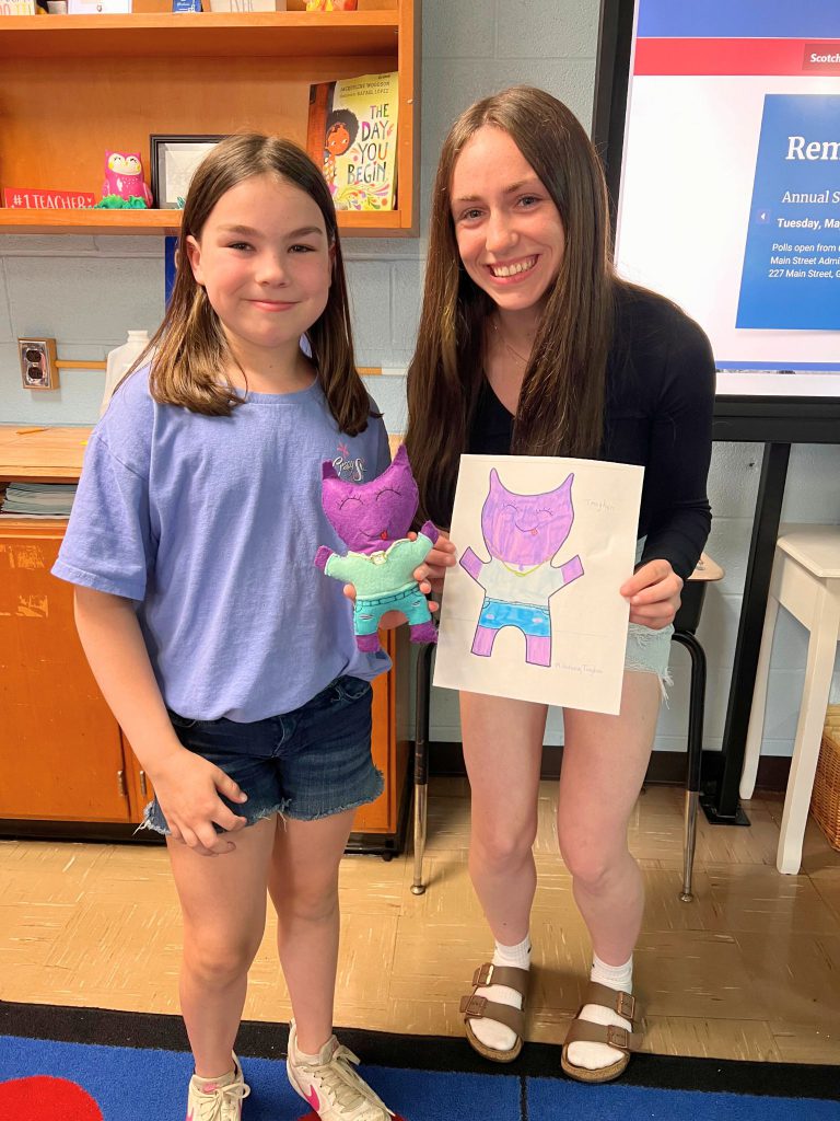 A third grade student stands to the left of a high school student. The young student is holding the sewn monster from the drawing that the older student is holding. The monster is purple and has a t-shirt on and shorts.