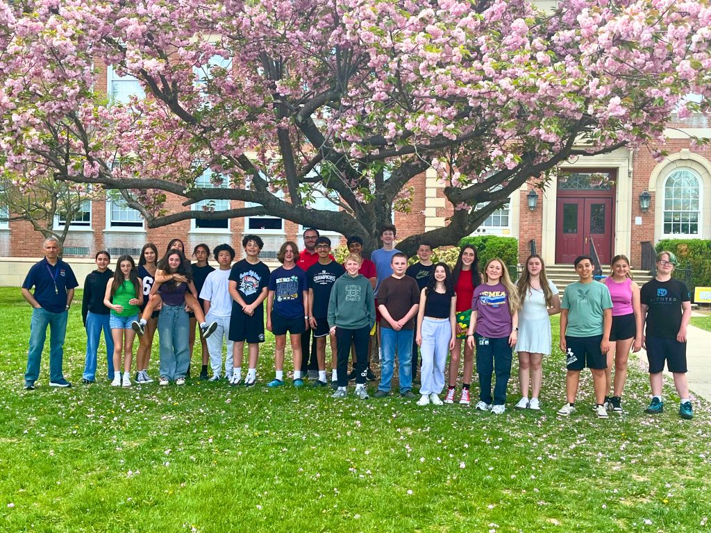 Students in C. J. Hooker Middle School are standing in the front yard of the school and smiling for the camera. They are under a tree with pink, white and green leaves and flowers.