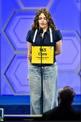 Cora Massaro stands at the microphone at the Scripps National Spelling Bee. There is a judge in the foreground looking at her. She has number 143 on the card she is wearing and her name, along with Orange-UIster BOCES.
