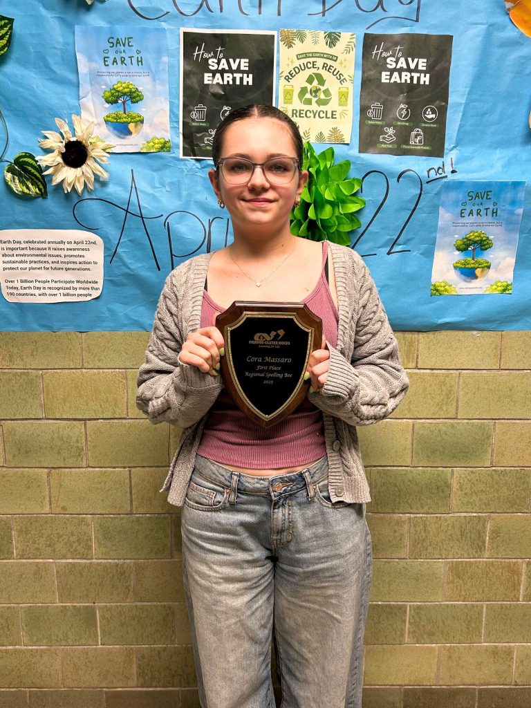 Cora Massaro stands in front of a wall celebrating Earth Day and holds a plaque with her name on it from Orange-Ulster BOCES recognizing her Spelling Bee win.