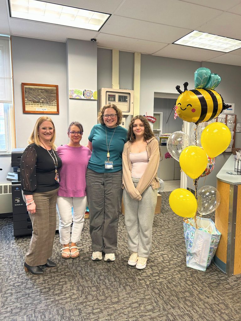 Three administrators and Cora Massaro stand in an office with Bee themed balloons and a gift bag as part of Cora's send-off to the Scripps National Spelling Bee.