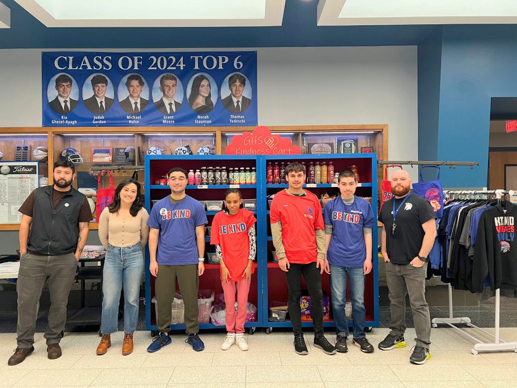 Seven people stand next to each other for a photo at the G.H.S. Kindness Cart. The four students in the middle are wearing shirts that say "Be Kind" and the staff are standing on both sides of the students and are smiling for the camera.