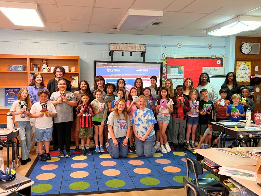 All the students and teachers involved in the monsters sewing project are smiling for a photo. The younger students are holding up their monsters in front of the older students behind them.