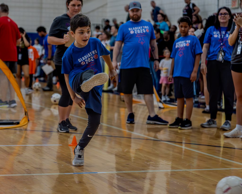 A Goshen student kicked a soccer ball with coaches and athletes looking on during a competition during the Special Olympics in Goshen High School. His leg is in the air after the kick and he is watching the ball travel toward the goal.