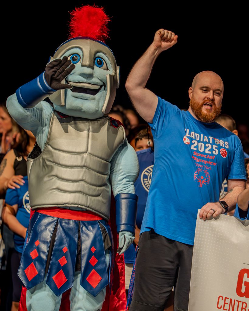 Goshen Special Olympics coach raises his hand in celebration of the opening ceremony next to the Goshen Gladiator on stage.