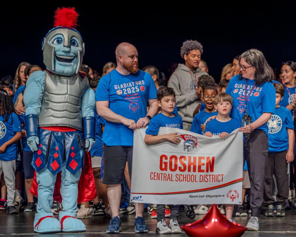 The Goshen Central School District stands on stage with their Special Olympics sign. The Gladiators mascot is next to them to the left.