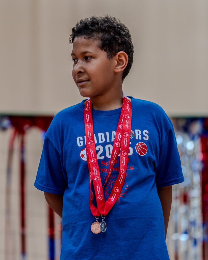 A Goshen student stands on a platform with medals on and looks off to the left. He has two medals and is wearing a Goshen Central School District shirt.