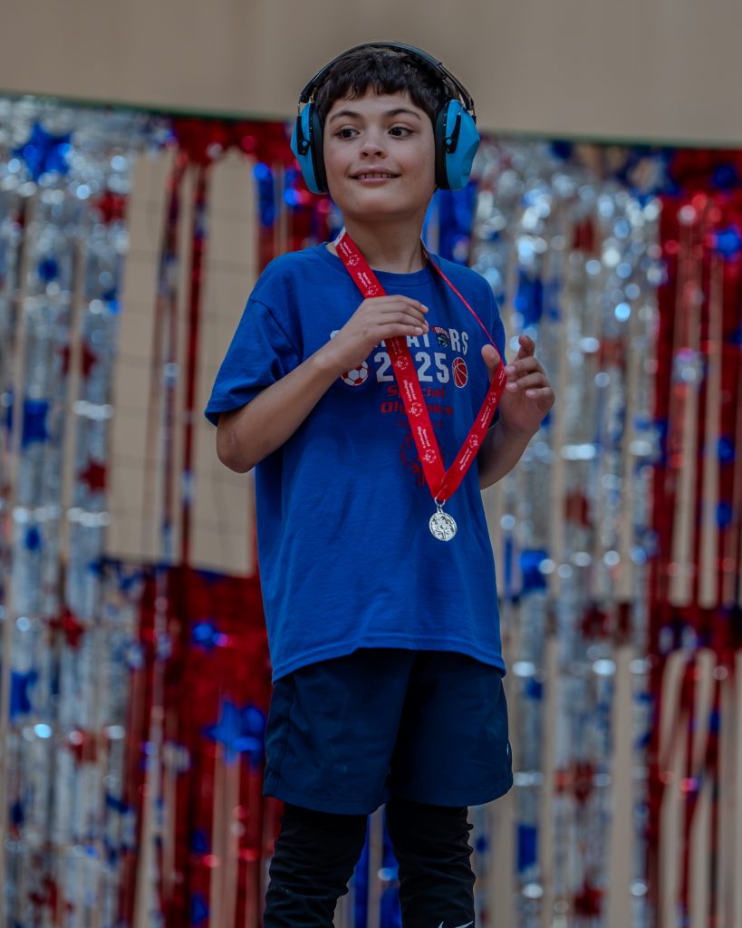 A Goshen student stands on stage and holds a medal. He is wearing a Goshen shirt and smiling out at the crowd.