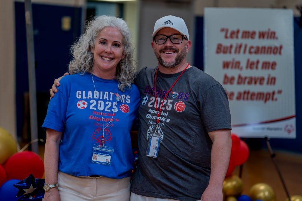 Two Goshen staff smile and look at the camera with Gladiators 2025 Special OIympics shirts. They are both wearing lanyards with shirts that have the Special Olympics logo on them as well.
