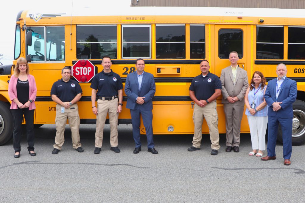 Eight people stand next to a bus with their arms crossed and smile. They are all dressed nicely for the photo.
