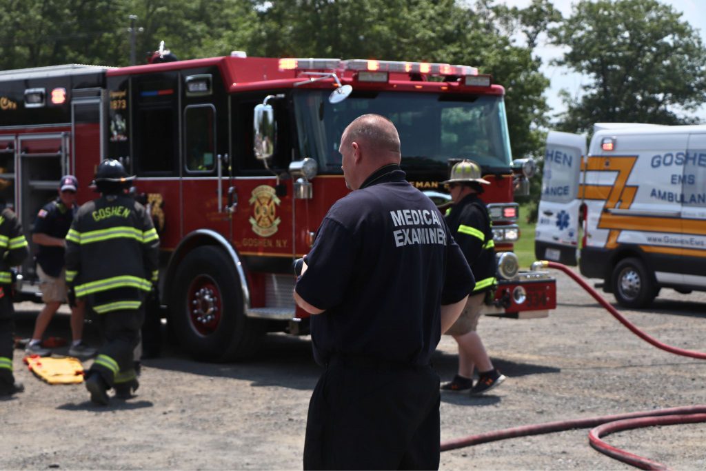 The medical examiner is watching the scene play out while they take photos of the events from the crash. The fire truck and ambulance is in the distance.