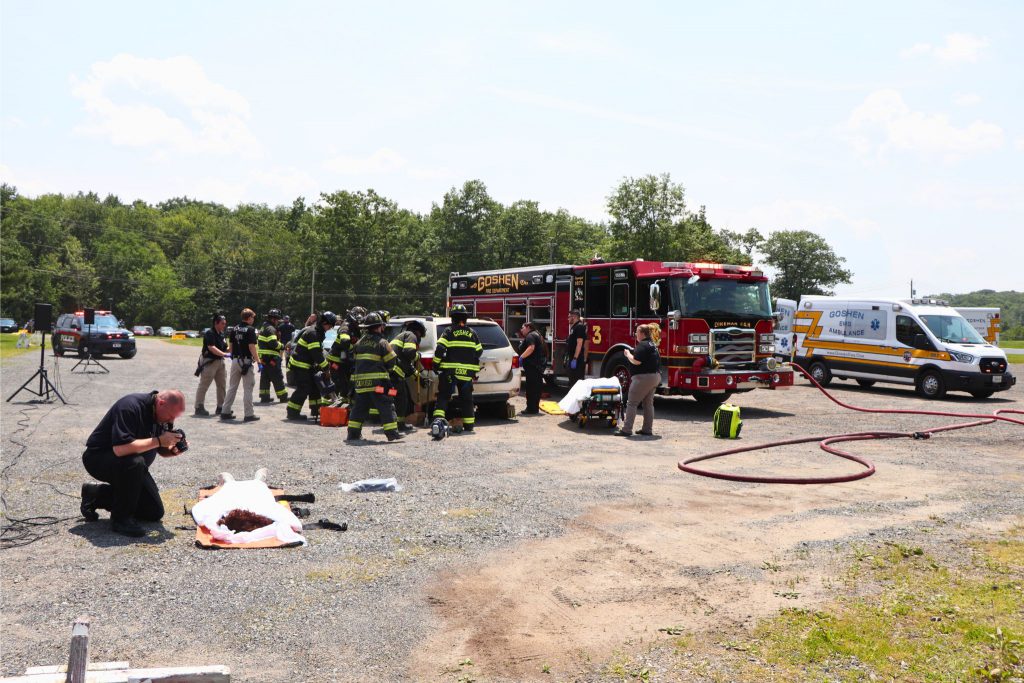 A medical examiner is taking photos of a dummy on the ground while emergency personnel are in the background attending to a crashed car.
