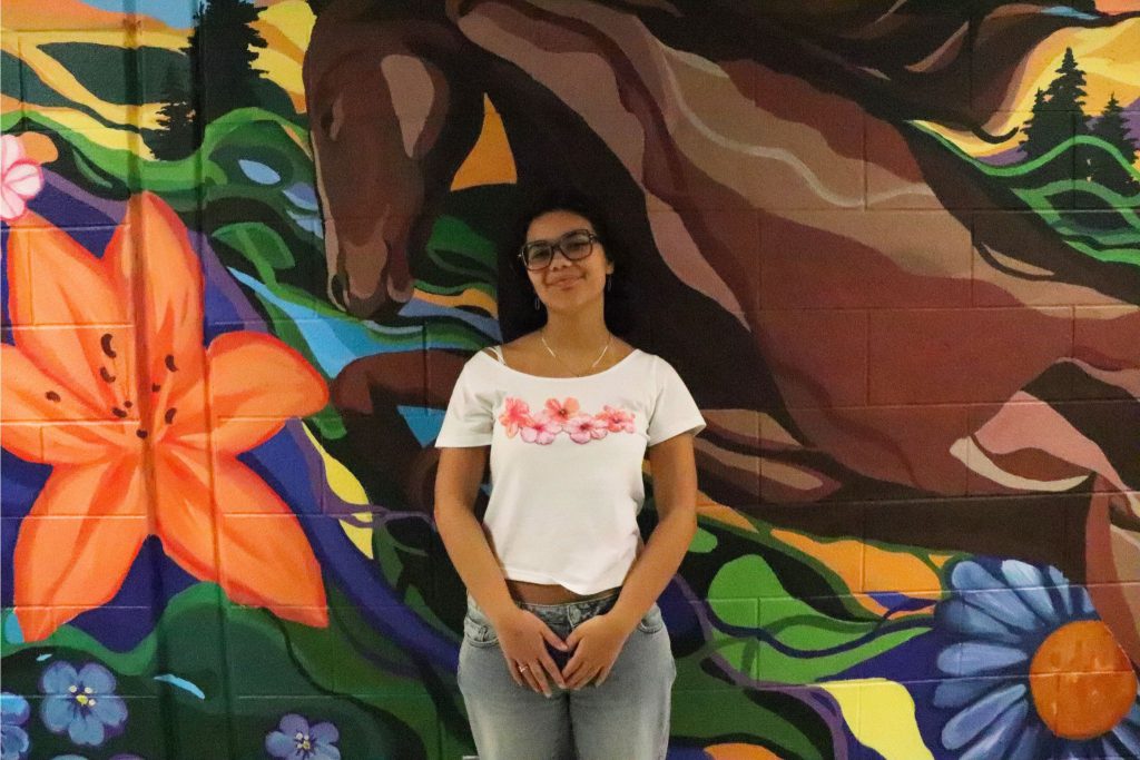 Katie Palau stands in front of the mural she created on the wall in Goshen High School and smiles. The horse she painted is behind her.