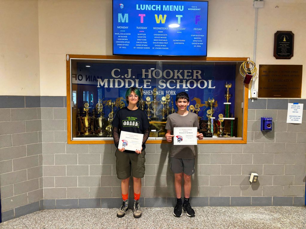 Two students are holding up their student of the month certificates in the hallway. They are looking at the camera and smiling.