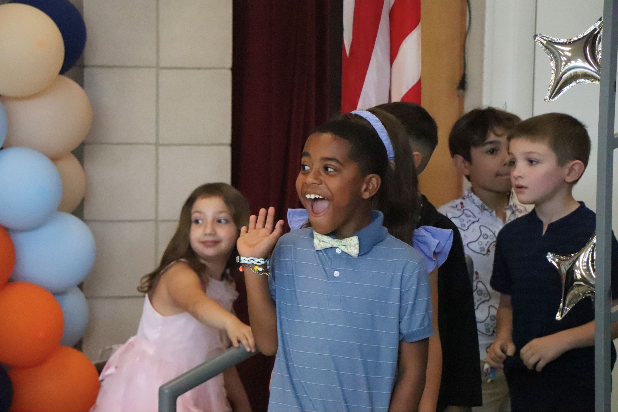A student smiles and waves to the camera while wearing a bowtie and a collared shirt walking on stage. 