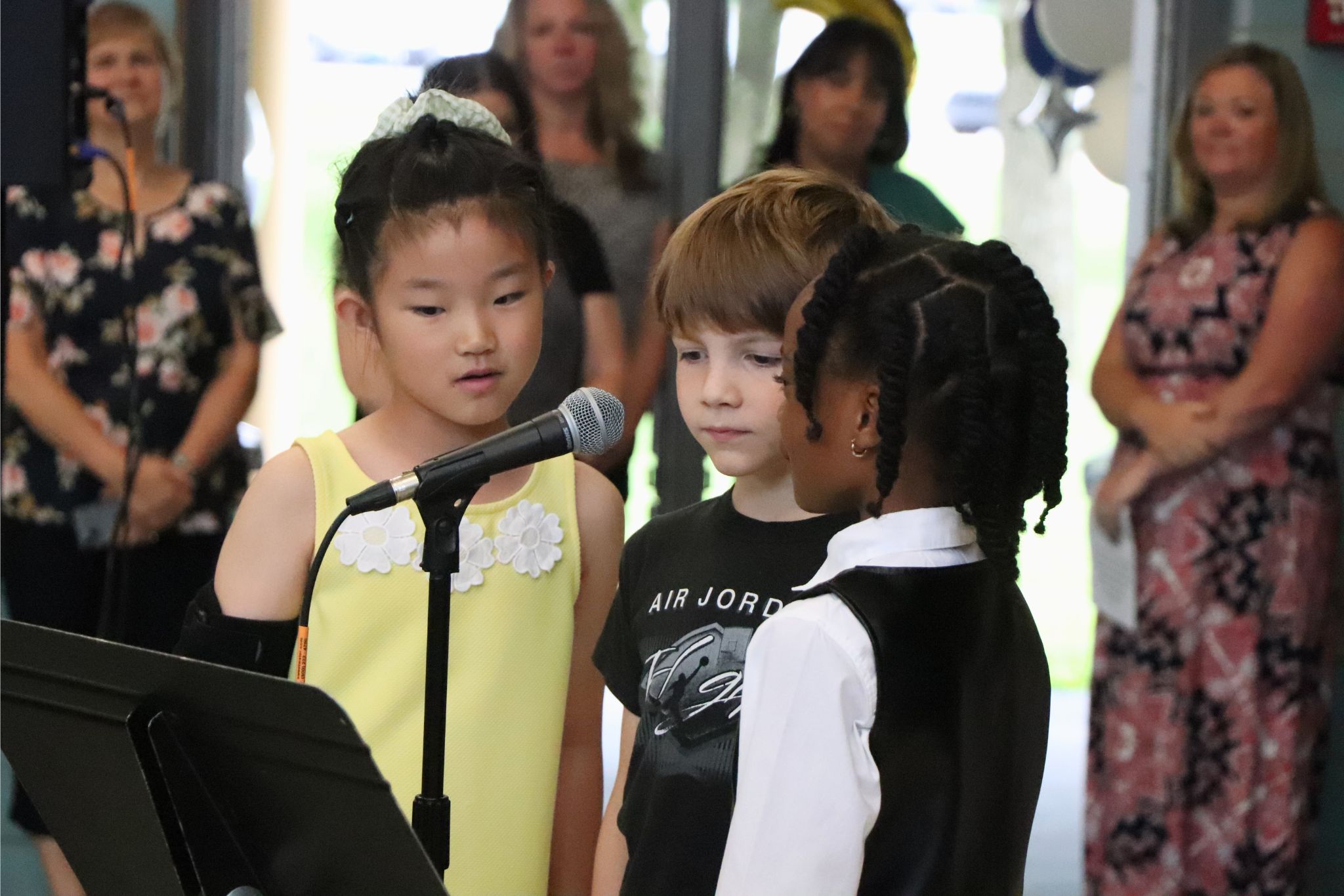 Three students stand at a microphone at the second grade moving up ceremony. 