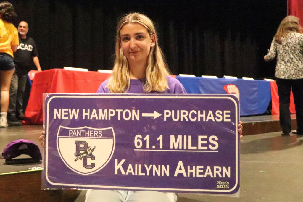 A student sits on the stage and holds up a sign that shows the distance they will be traveling to go to college and the logo of their institution.