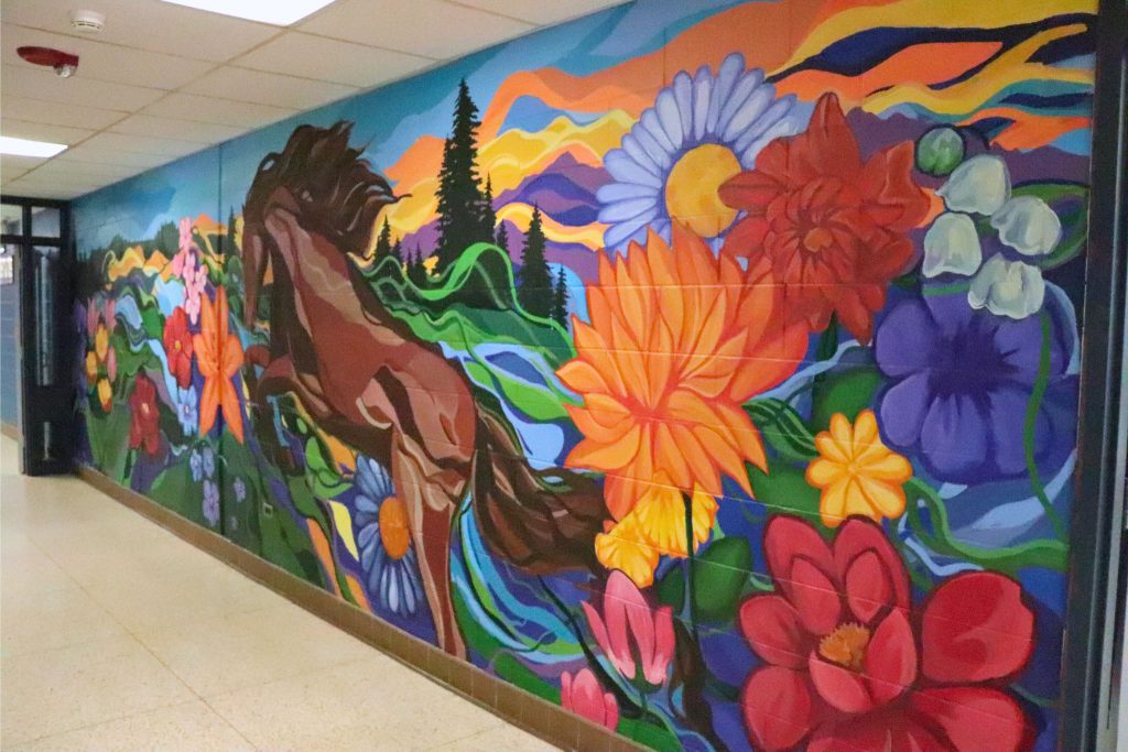 A finished side angle of the wall in Goshen High School. There are flowers and hills and trees and a horse on the wall.
