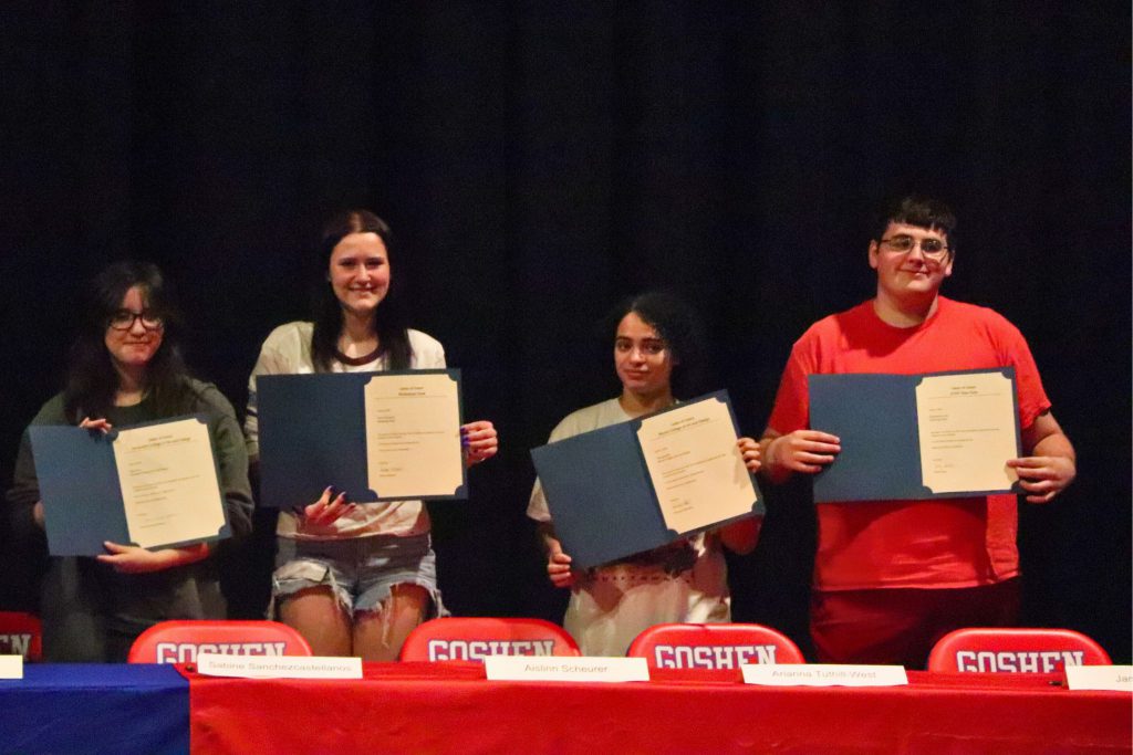 Four students at the end of the table smile for a photo at Unified Arts Signing Day. They are all looking at the camera and holding up their letters.