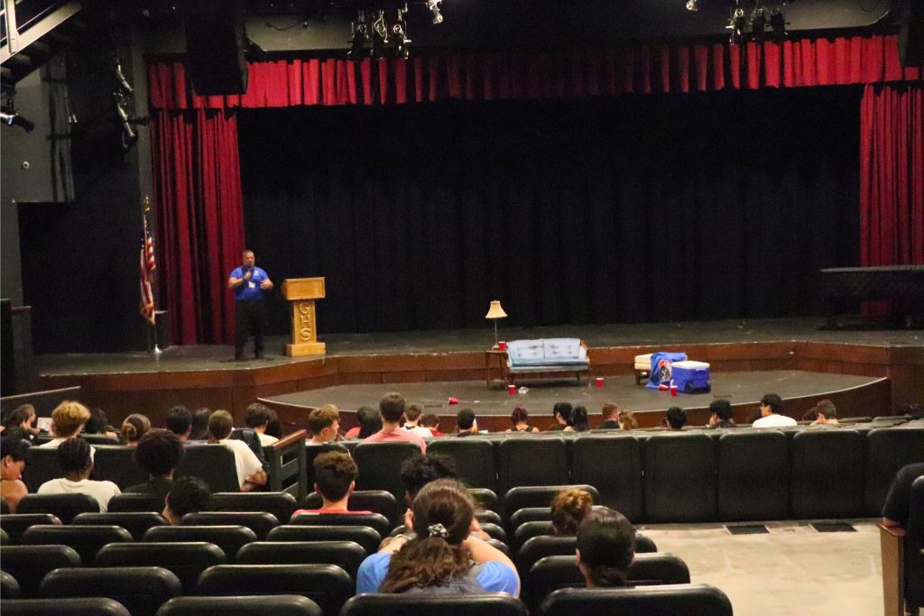 The principal is speaking on stage with students in the crowd of the auditorium.