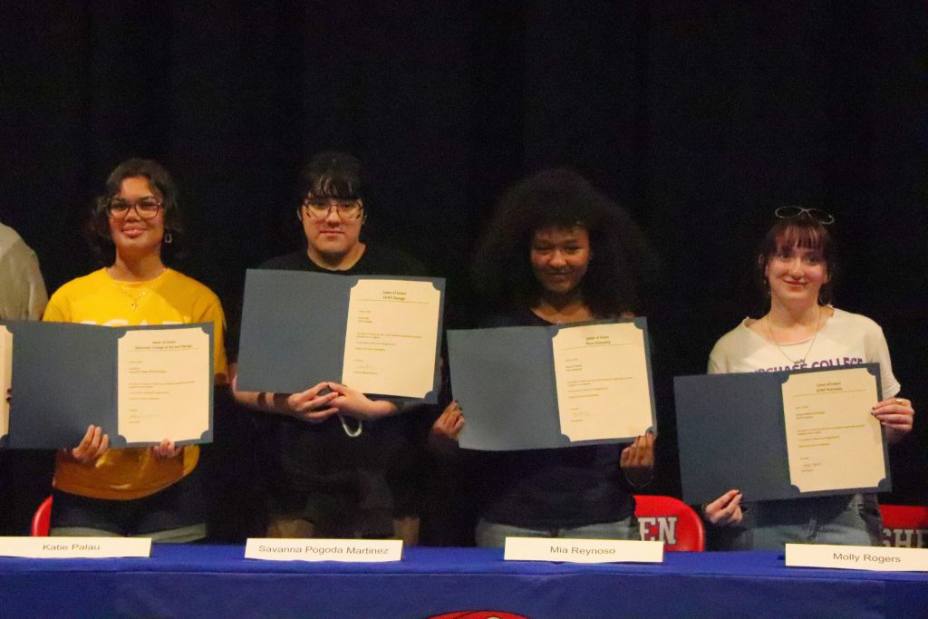Four students smile for a photo centered behind the table at Unified Arts Signing Day. They are all looking at the camera and holding up their letters.