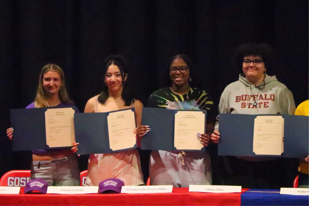 Four students smile for a photo behind the table at Unified Arts Signing Day. They are all looking at the camera and holding up their letters.